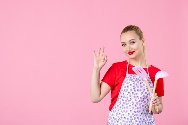 Вид спереди молодая домохозяйка держит столовые приборы на розовой стене