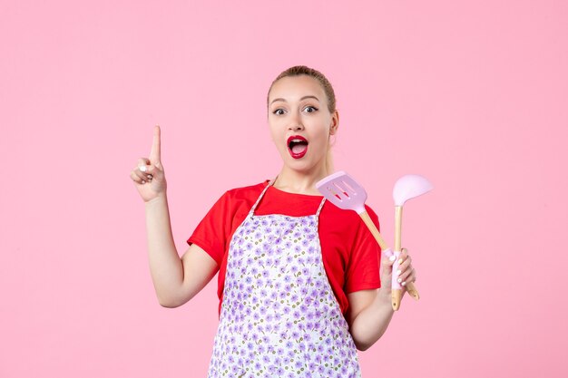 Вид спереди молодая домохозяйка позирует со столовыми приборами в руках на розовой стене