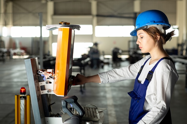 A front view young lady in blue construction suit and helmet controlling machines in hangar during daytime buildings architecture construction