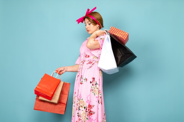 A front view young lady in flower designed pink dress posing holding shopping packages on blue