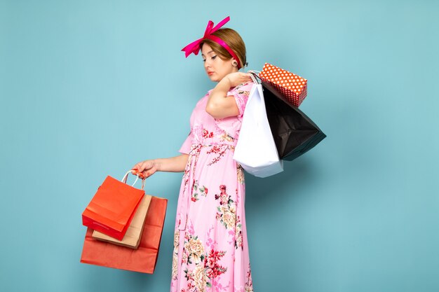 A front view young lady in flower designed pink dress posing holding shopping packages on blue