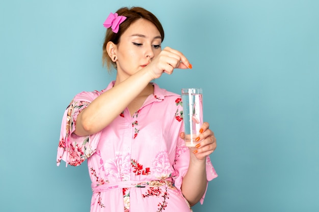 Молодая дама в розовом платье, вид спереди, кладет таблетку в стакан с водой на синем