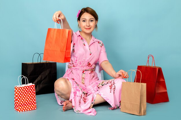 A front view young lady in flower designed pink dress sitting and posing with smile and shopping packages on blue