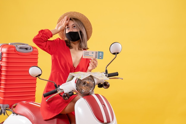 Front view young lady on moped with red suitcase holding ticket putting hand on her forehead