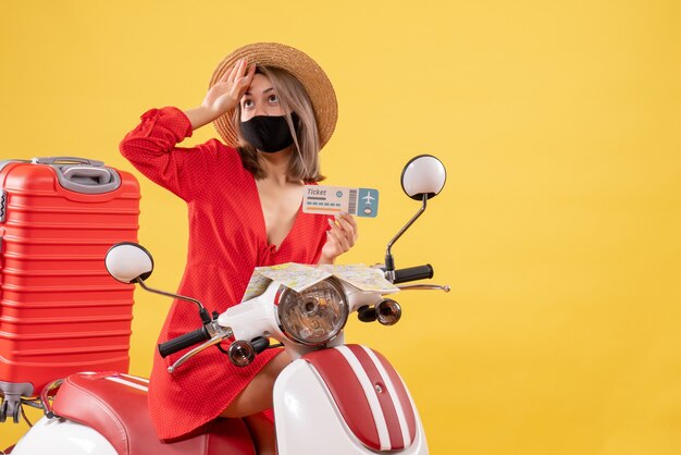 Front view young lady on moped with red suitcase holding ticket putting hand on her forehead