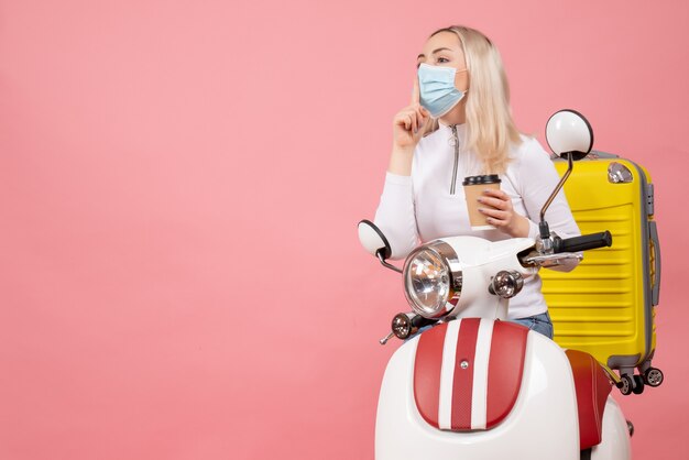 Front view young lady on moped with yellow suitcase holding coffee cup making hush sign