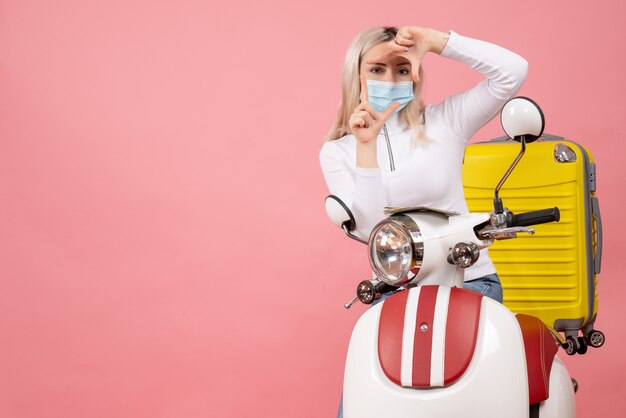 Front view young lady on moped with yellow suitcase making camera sign