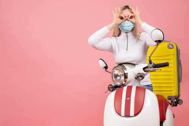 Front view young lady on moped with yellow suitcase making hands binoculars