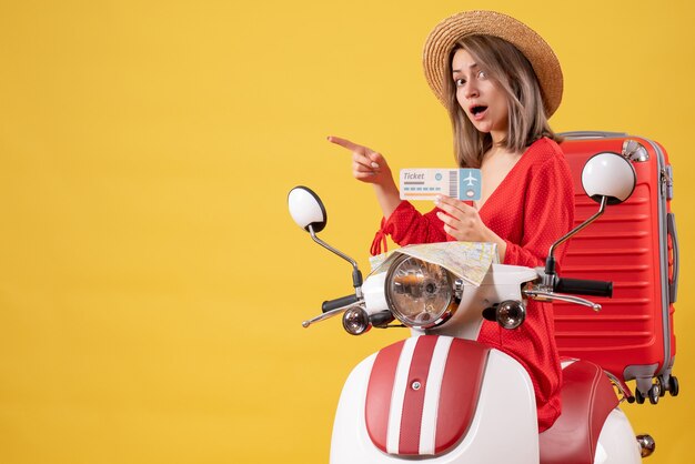 Front view young lady in red dress holding ticket pointing at left on moped