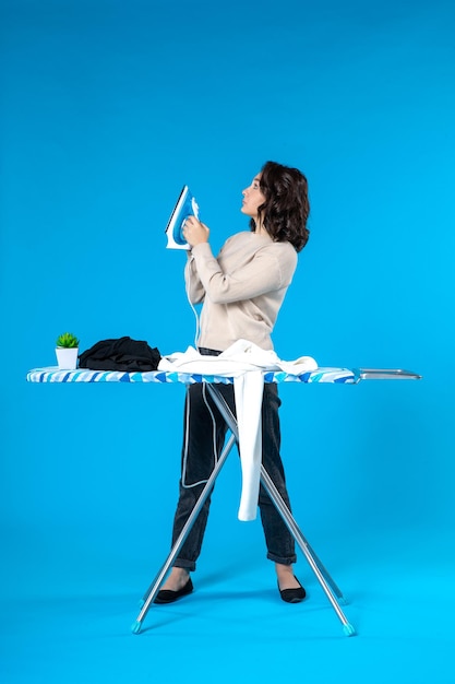 Front view of young lady standing behind the board and ironing the clothes on blue surface