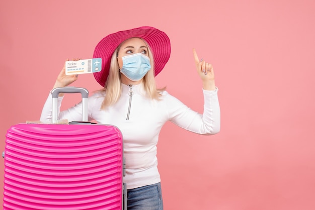 Front view young lady standing near suitcase holding plane ticket pointing at ceiling