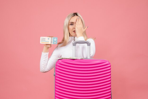 Front view young lady standing behind pink suitcase holding ticket putting hand on her eye