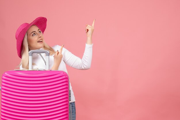 Front view young lady standing behind pink suitcase pointing finger up