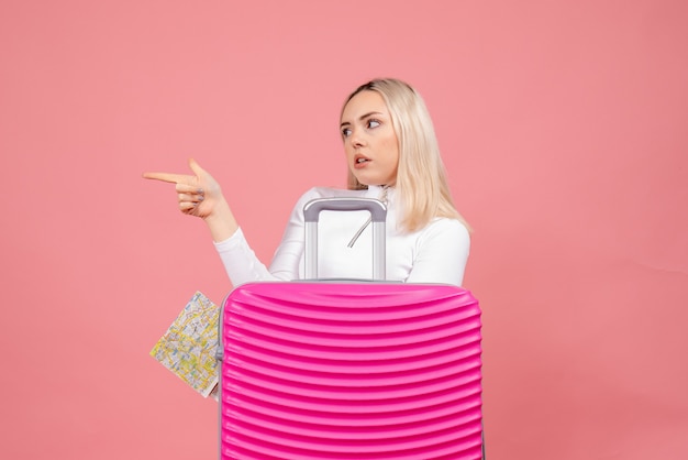 Front view young lady standing behind pink suitcase pointing at left holding map