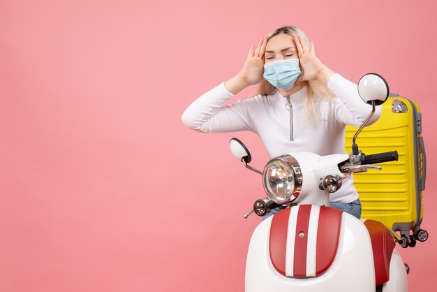 Front view young lady with closed eyes on moped with yellow suitcase holding her head