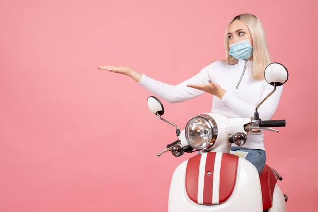 Front view young lady with mask pointing at left standing near moped