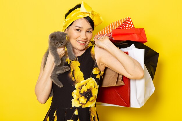 Вид спереди молодой леди в желто-черном цветочном платье с желтой повязкой на голове, держащей пакеты с покупками и котенка на желтом