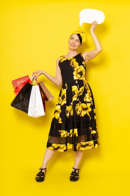 A front view young lady in yellow-black flower designed dress with yellow bandage on head holding shopping packages white sign on the yellow