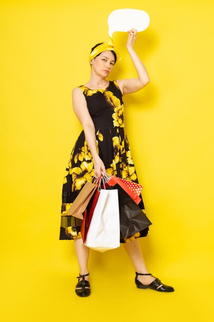 黄色の黒い花の正面図の若い女性は黄色のショッピングパッケージ白い看板を保持している頭の上の黄色の包帯でドレスを設計しました