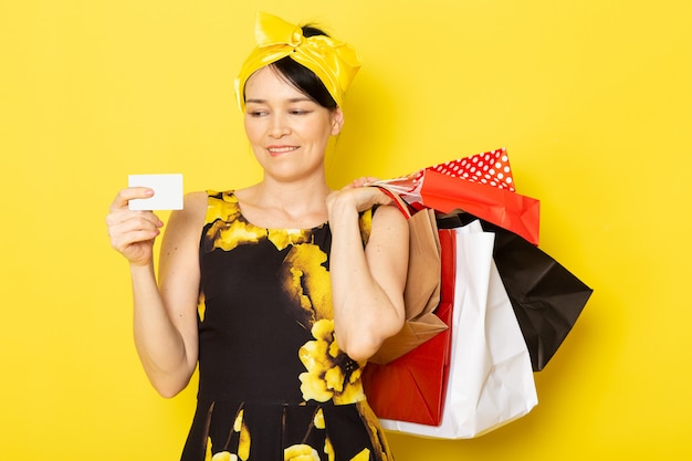 Вид спереди молодой леди в желто-черном цветочном платье с желтой повязкой на голове, держащей пакеты с покупками на желтом