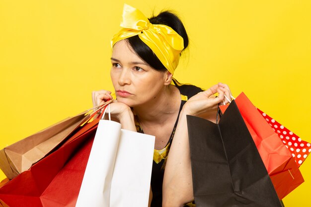 A front view young lady in yellow-black flower designed dress with yellow bandage on head thinking holding shopping packages on the yellow