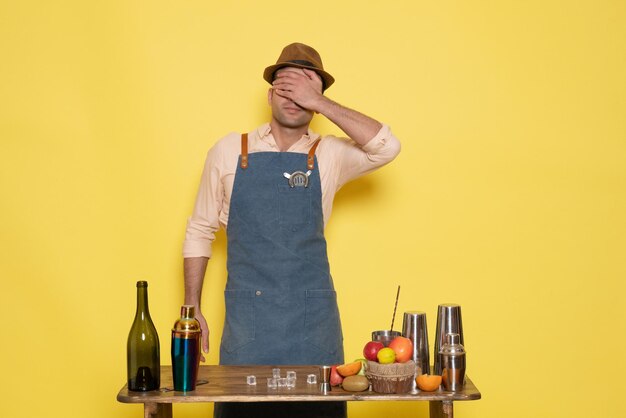 Front view young male bartender in front of table with drinks and shakers on yellow background