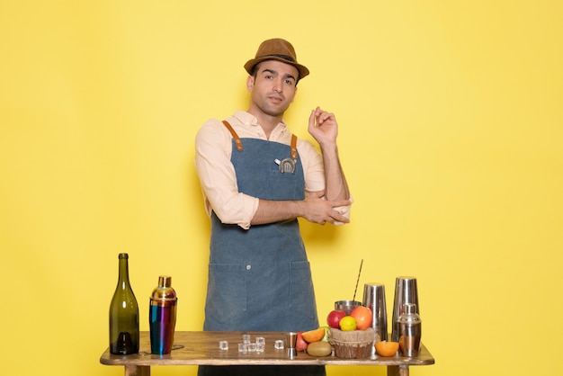 Front view young male bartender in front of table with shakers and drinks on the yellow background