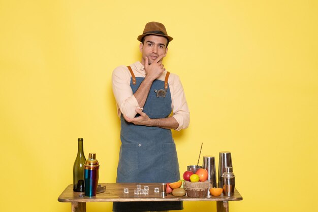 Front view young male bartender in front of table with shakers and drinks on yellow background