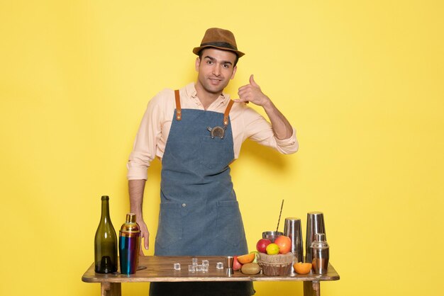 黄色の背景にシェーカードリンクとテーブルの前で正面図若い男性バーテンダー