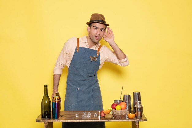 Front view young male bartender in front of table with shakers drinks on a yellow background
