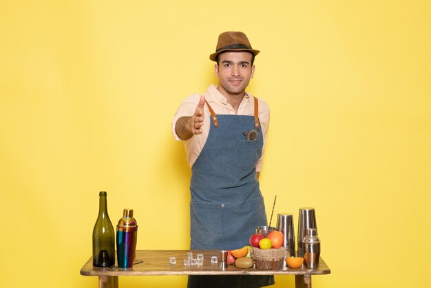 Front view young male bartender in front of table with shakers drinks on the yellow background