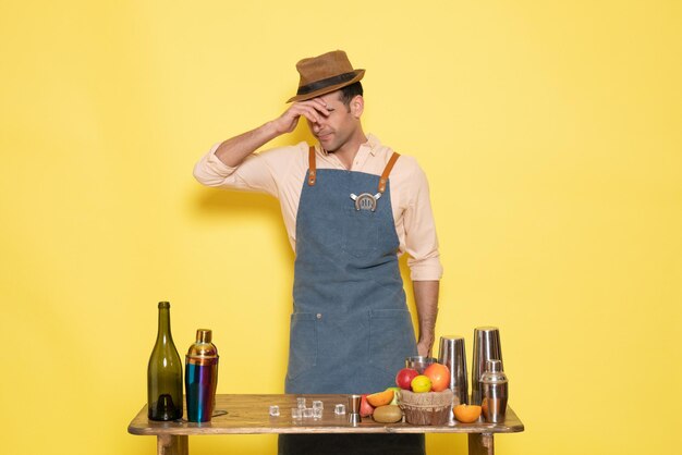 Front view young male bartender in front of table with shakers drinks on yellow background