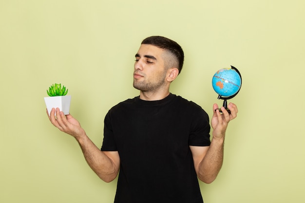 Вид спереди молодой самец в черной футболке держит маленькое зеленое растение и маленький глобус на зеленом