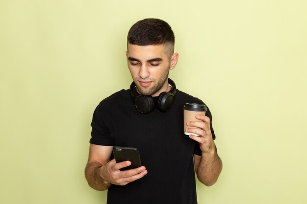 携帯電話を押しながら緑のコーヒーカップを保持している音楽を聴く黒いtシャツで正面の若い男性