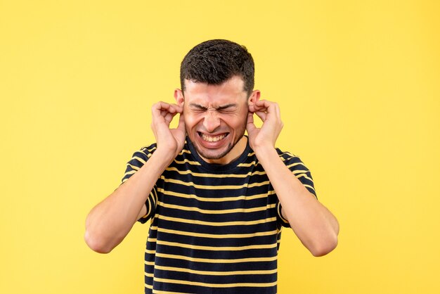 Front view young male in black and white striped t-shirt closing his ears with hands on yellow isolated background