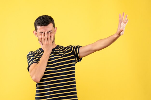 Front view young male in black and white striped t-shirt covering face with hand on yellow isolated background