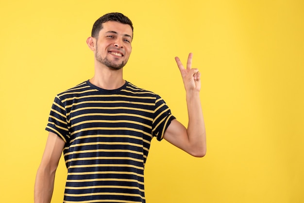 Front view young male in black and white striped t-shirt making victory sign on yellow isolated background