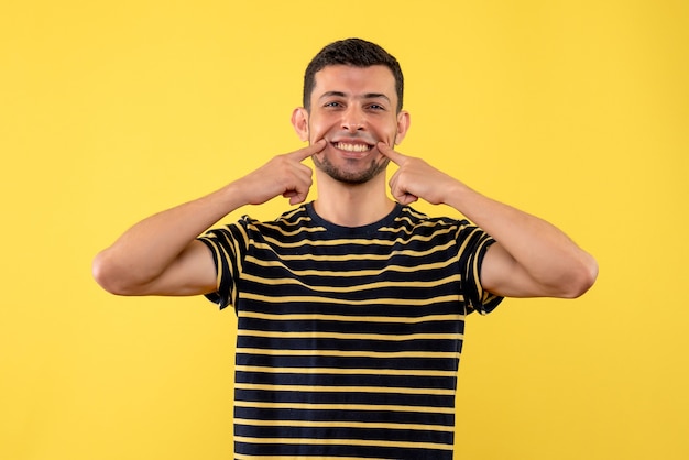 Front view young male in black and white striped t-shirt pointing at his smile on yellow isolated background