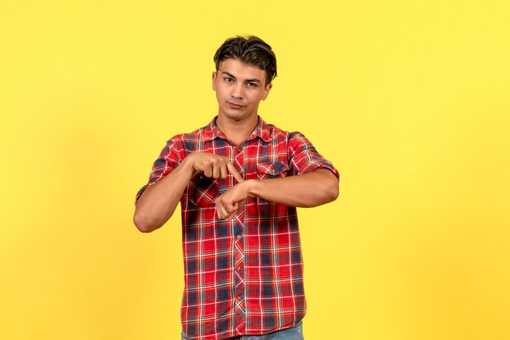 मुफ़्त फोटो सामने से पीले डेस्क पर अपनी कलाई की ओर इशारा करते हुए उज्ज्वल शर्ट में युवा पुरुष मॉडल।