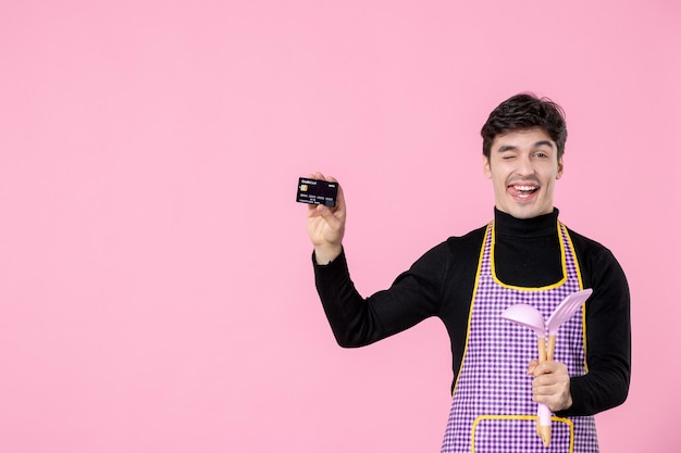 Vista frontale giovane maschio in mantello che tiene carta bancaria e cucchiai su sfondo rosa lavoro lavoratore colore denaro capo cucina orizzontale cucina uniforme professione