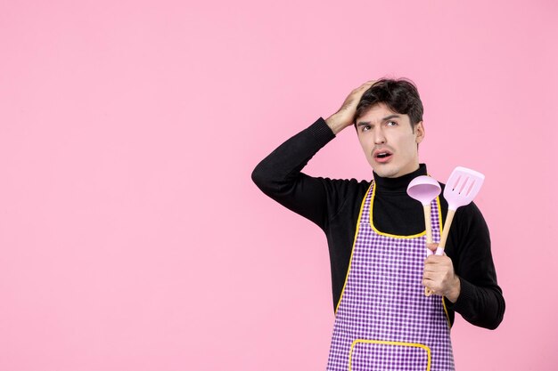Вид спереди молодой мужчина в плаще держит розовые ложки на розовом фоне тесто профессия кухня униформа цвет горизонтальный главный кулинар работники