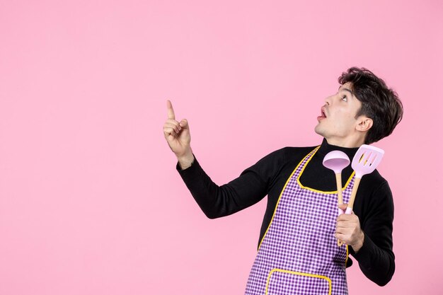 Front view young male in cape holding pink spoons on pink background dough profession cuisine uniform horizontal chief cooking job color worker