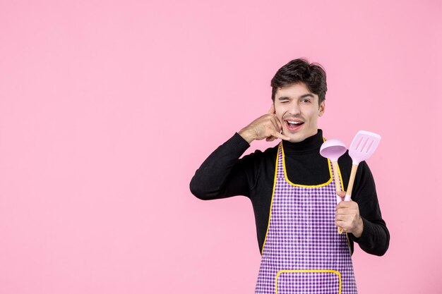 Front view young male in cape holding pink spoons on pink background dough profession cuisine work uniform color horizontal cooking cook
