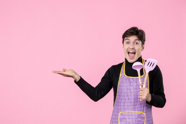 Front view young male in cape holding pink spoons on pink background profession cuisine work dough uniform horizontal cooking