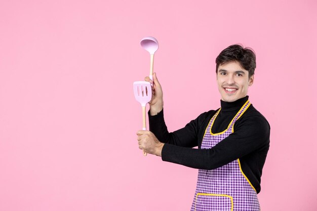 Front view young male in cape holding spoons on pink background job dough profession cuisine cooking worker chief color horizontal uniform