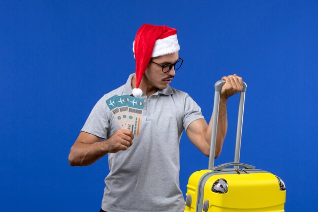 青い壁の飛行休暇の飛行機のチケットと重いバッグを運ぶ正面図若い男性