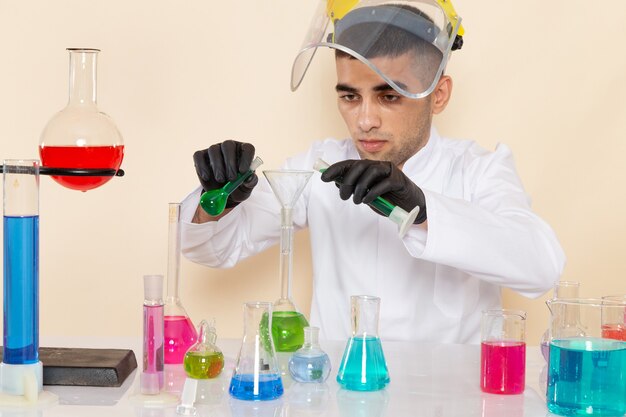 Front view young male chemist in white special suit in front of table with colored solutions working with them on the cream desk lab science work chemicals