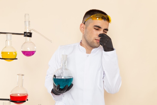 플라스크를 들고 크림 책상 실험실 화학 과학에 그의 코를 닫는 솔루션 테이블 앞의 흰색 특수 정장에 전면보기 젊은 남성 화학자