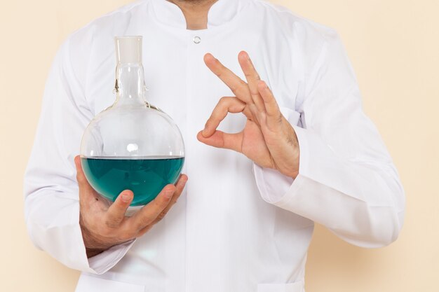 クリーム色の壁の科学実験化学実験室科学の青い溶液とフラスコを保持している白い特別なスーツの正面図若い男性化学者