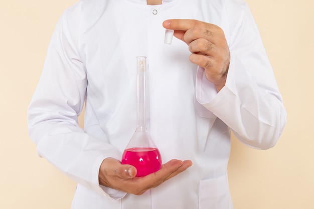 Вид спереди молодой химик-мужчина в белом специальном костюме, держащий колбы с розовым раствором на кремовой стене, научная лаборатория, эксперимент, химия, научная.